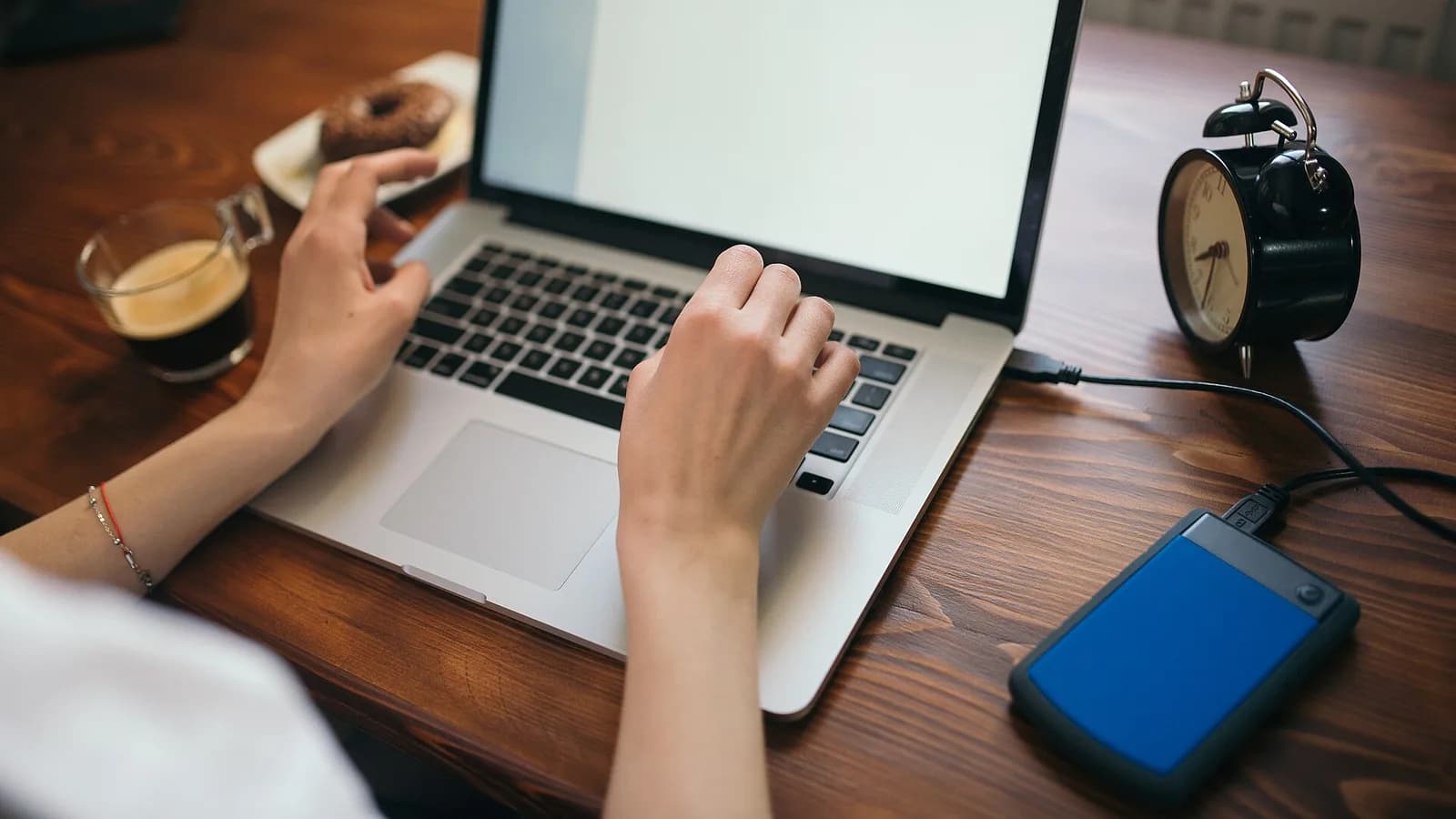 A person typing on a silver MacBook at a dark wooden desk, with a blue portable external hard drive plugged into it by a USB cable, beside a glass cup of coffee and a small black alarm clock