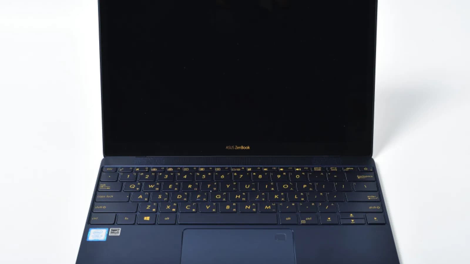 Komputer riba ASUS ZenBook biru gelap terbuka di atas permukaan putih dengan skrinnya kosong dan dimatikan, menampakkan papan kekunci hitam berhuruf emas, pembaca cap jari di penjuru trackpad dan pelekat kecil Intel Core i5