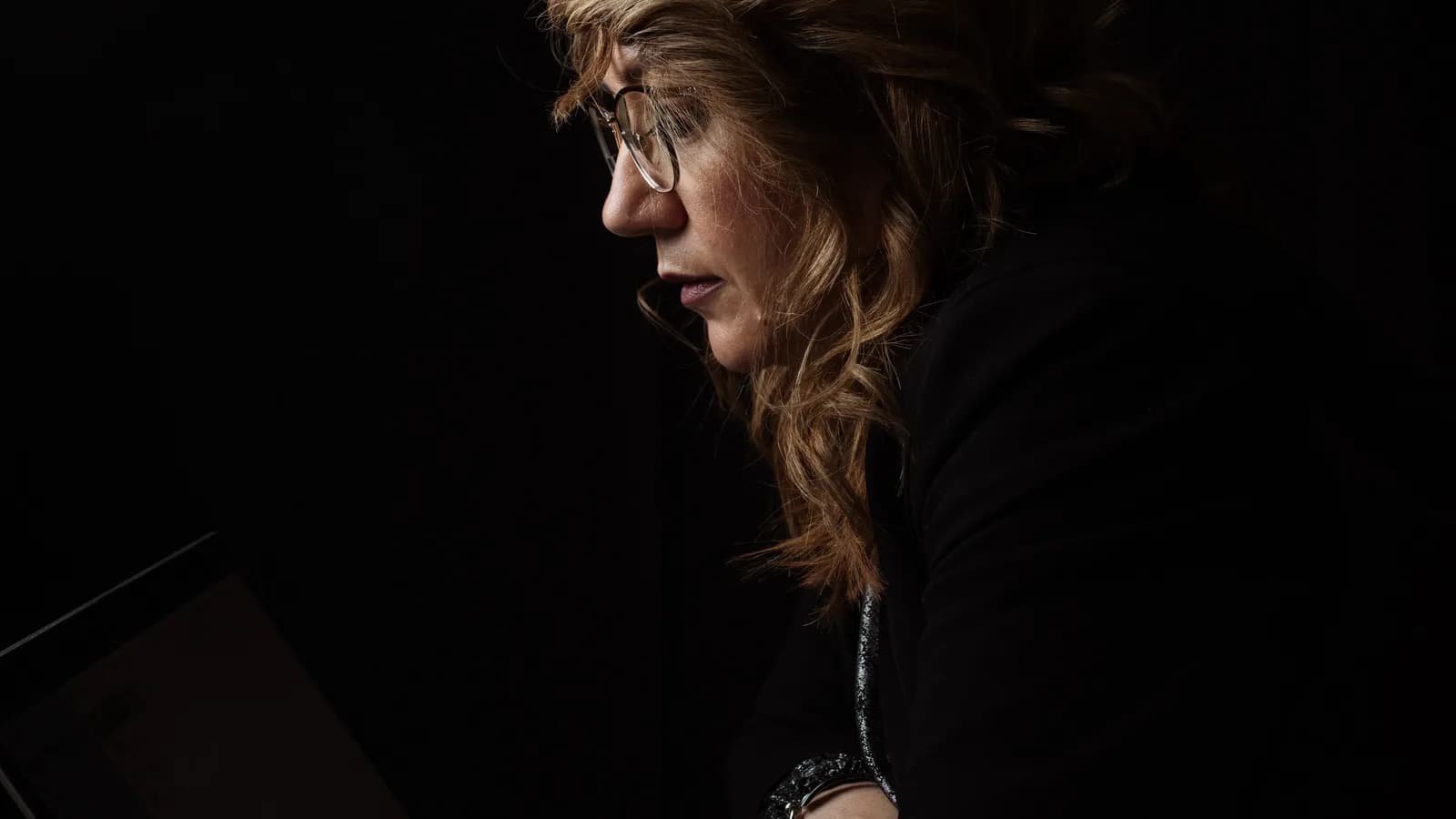 A woman in glasses seen in profile in a very dark room, her face and hair lit while a laptop screen glows faintly at the lower-left edge of the frame.