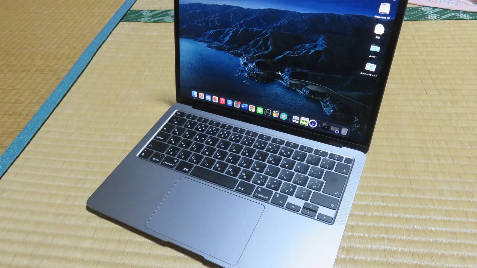 An open space-grey Apple laptop on a tatami mat, seen from the front left: its Japanese keyboard and wide trackpad, and a dark blue coastal wallpaper on the screen with a row of app icons along its bottom edge