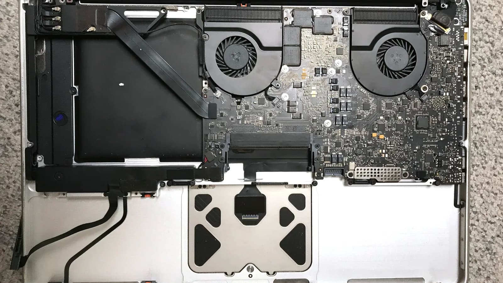 Bahagian dalam MacBook Pro yang dibuka dilihat dari bawah, penutup bawahnya ditanggalkan: papan litar dan dua kipas penyejuk di bahagian atas, sebuah kotak hitam besar di atas sebelah kiri, bahagian bawah trackpad di tengah, dan kabel hitam yang longgar di ruang kosong di bawah sebelah kiri