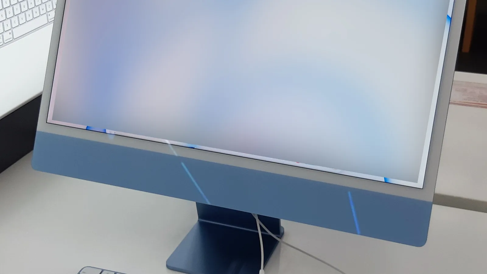 一台蓝色的一体机 iMac，从正面拍摄，放在浅色桌面上，纤薄的屏幕上显示着柔和、模糊的蓝粉色壁纸，下边框和扁平的支脚为同色的蓝色，旁边有一个白色键盘