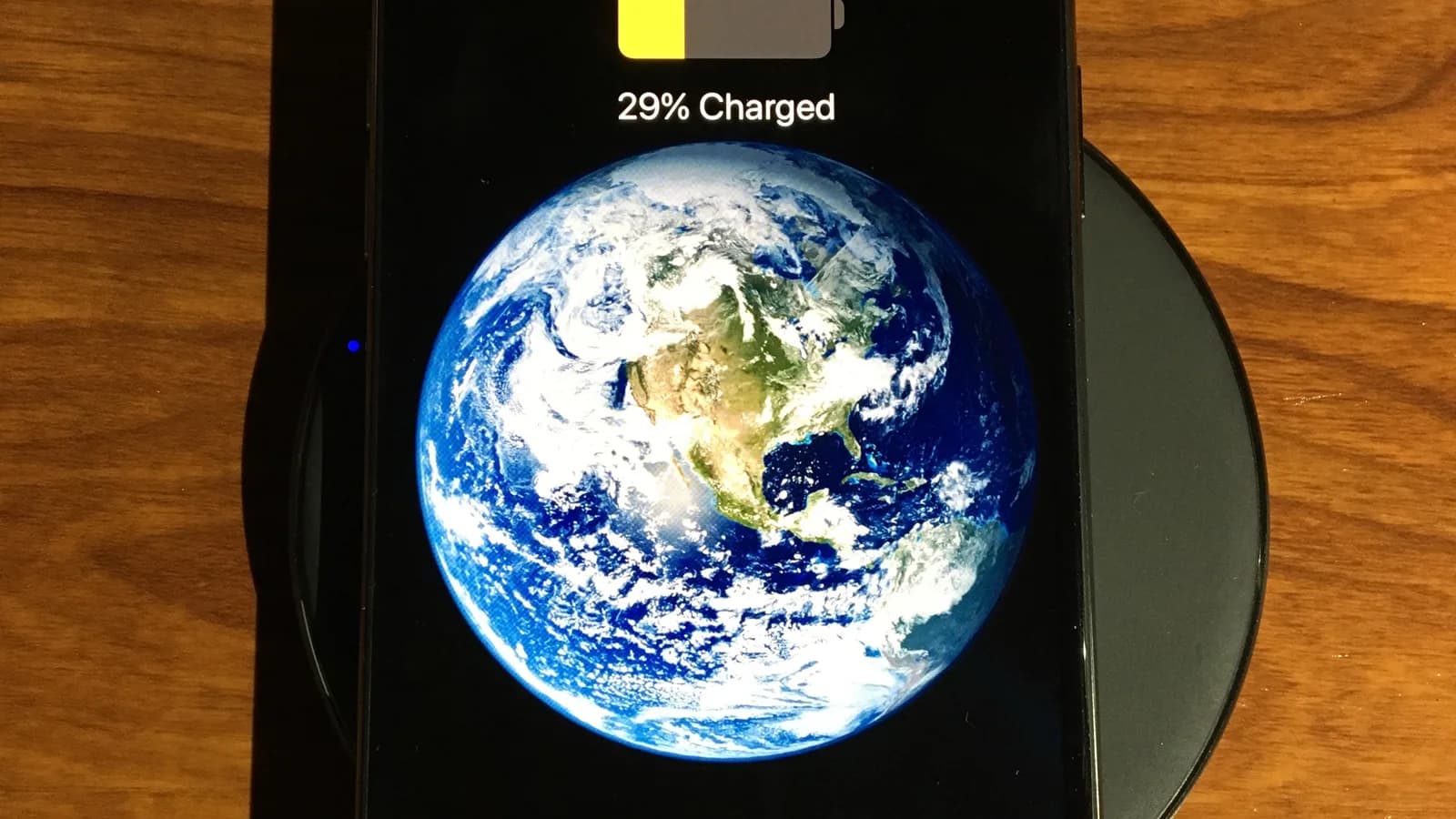 An iPhone face-up on a dark round wireless charging pad on a wooden table, its screen on and showing the Earth with the words "29% Charged" at the top