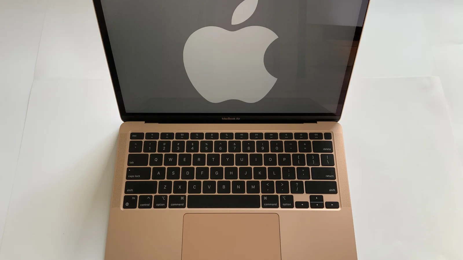 一台打开的金色 MacBook Air 笔记本电脑，从正前上方拍摄，放在白色台面上，屏幕上显示着一个大大的苹果标志，下方是黑色键盘和宽大的触控板