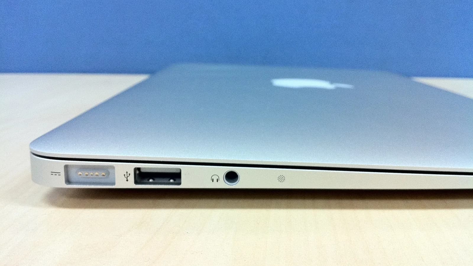 一台合上的银色 MacBook Air 的左侧，放在浅色木桌上，可见 MagSafe 电源口、USB-A 接口和耳机孔，顶盖上有苹果标志。
