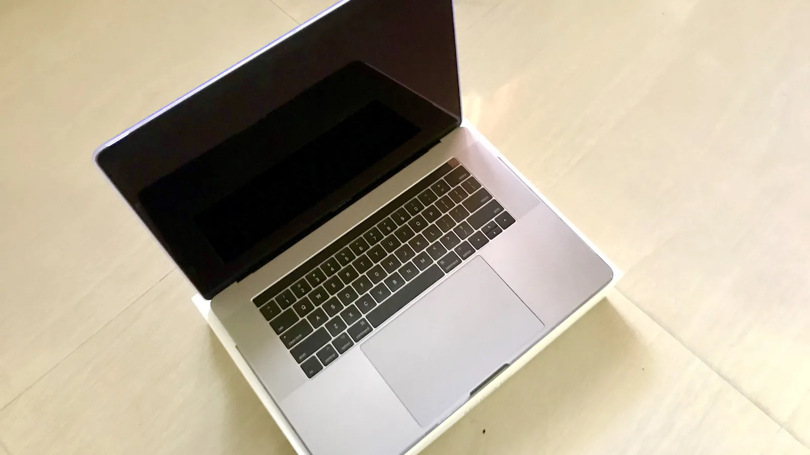 A space grey MacBook Pro open on top of its white box on a pale tiled floor, seen from above at an angle. The screen is off and dark; the black keyboard, the wide grey trackpad and the narrow Touch Bar strip along the top of the keys are all visible