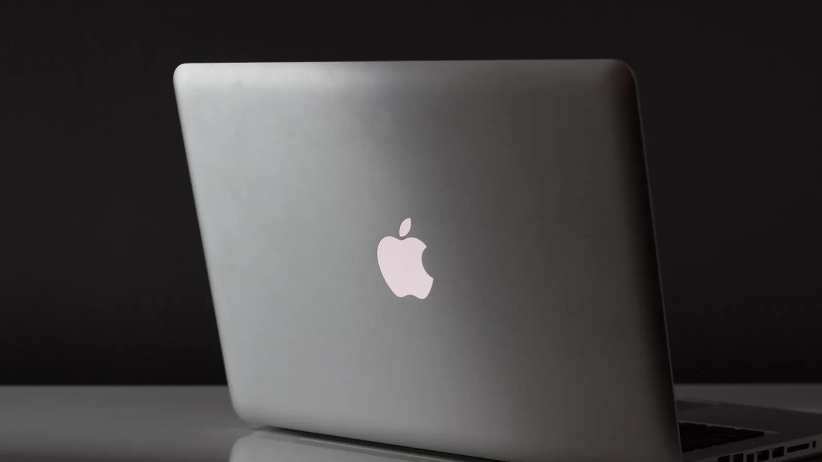 一台打开的银色铝制 MacBook 的背面，顶盖上发光的苹果标志亮着，背景昂暗，下方是反光的表面