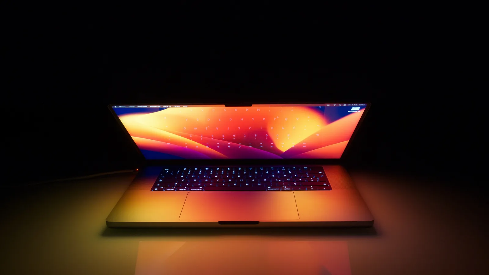 Sebuah komputer riba Apple yang terbuka di dalam bilik gelap, skrinnya bercahaya dengan kertas dinding jingga dan ungu yang menerangi papan kekunci berlampu latar, trackpad dan meja di hadapannya