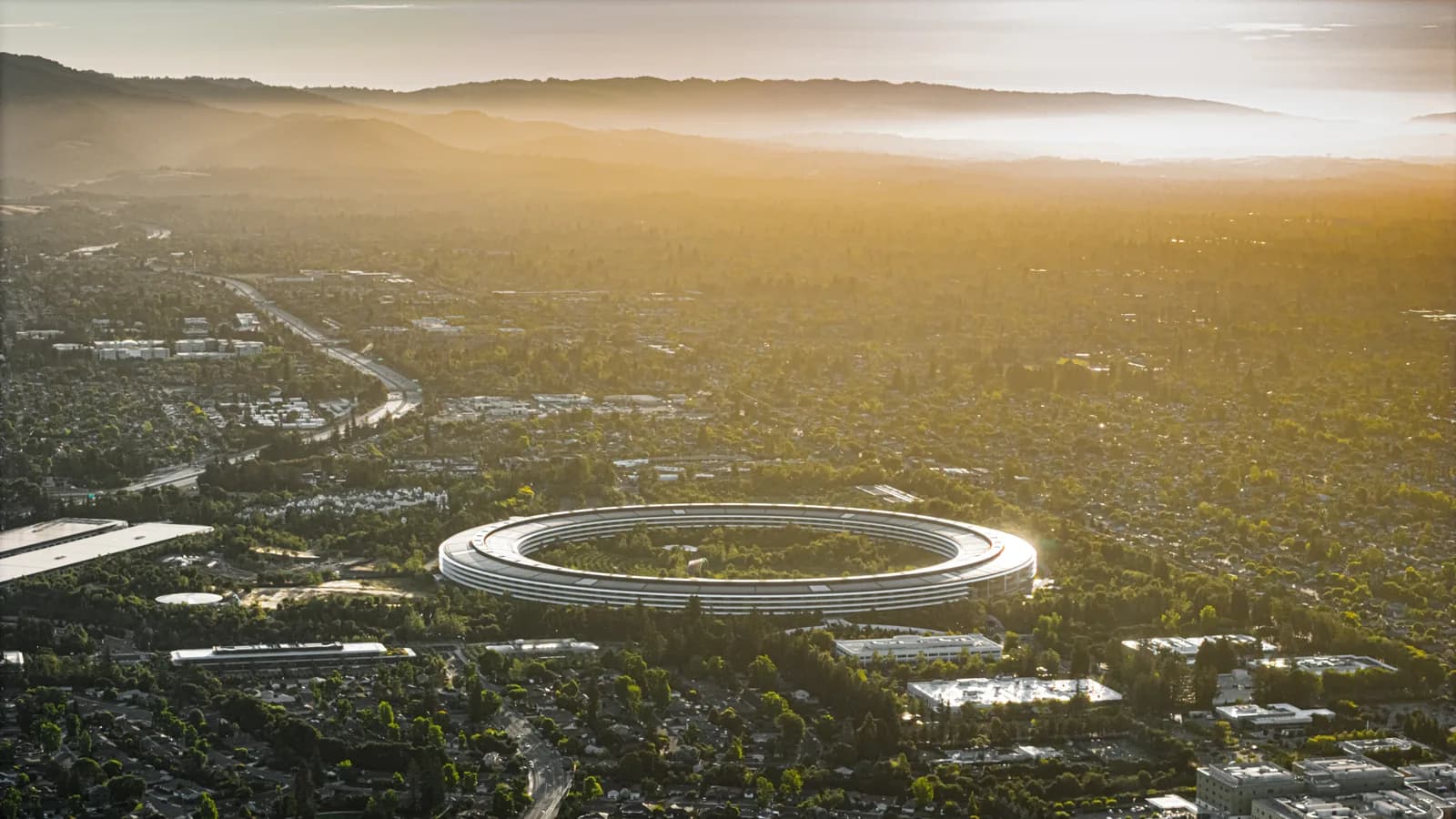 An aerial photograph at golden hour of Apple Park in Cupertino: the circular glass headquarters building sits inside a ring of trees in the middle of low suburban sprawl, with the ridgelines behind it fading into haze