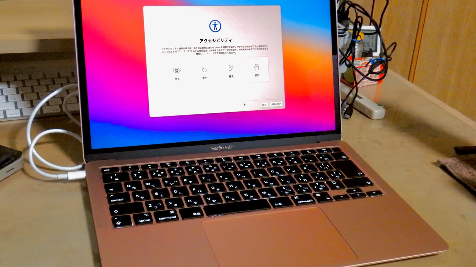 Sebuah MacBook Air emas yang terbuka di atas meja, skrinnya menunjukkan halaman Kebolehcapaian pembantu persediaan macOS, dengan kabel USB-C dipasang ke port kiri dan papan kekunci luaran di sebelahnya.