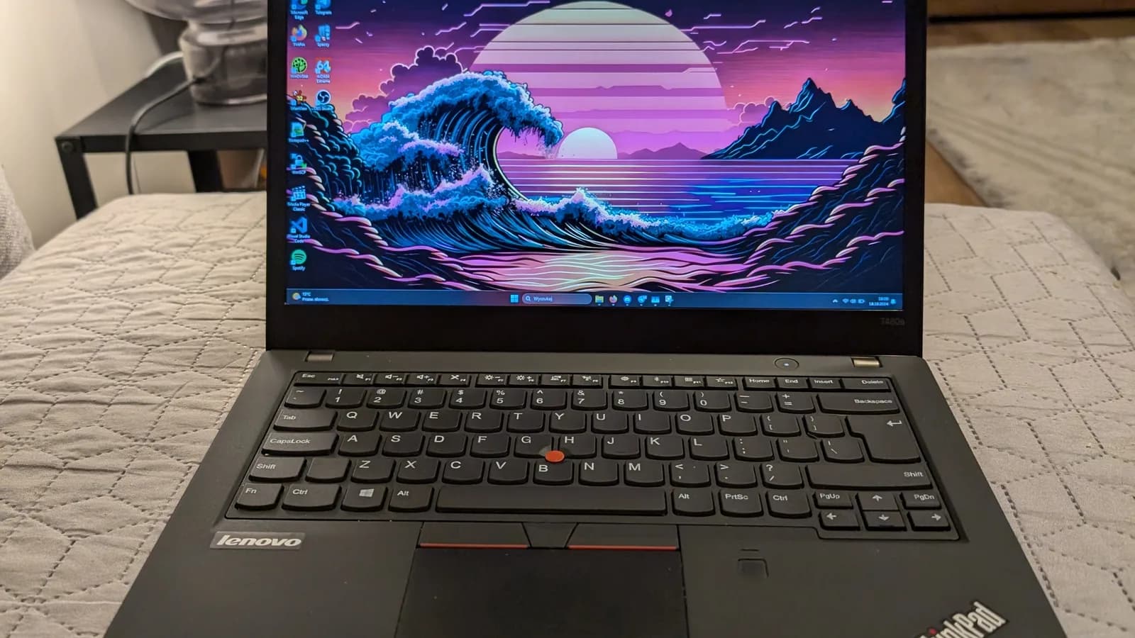 Sebuah komputer riba Lenovo ThinkPad hitam yang terbuka di atas cadar berjahit, skrinnya memaparkan kertas dinding ombak dan matahari terbenam dengan ikon desktop, dengan papan kekunci, kayu penuding merah dan butang trackpad kelihatan
