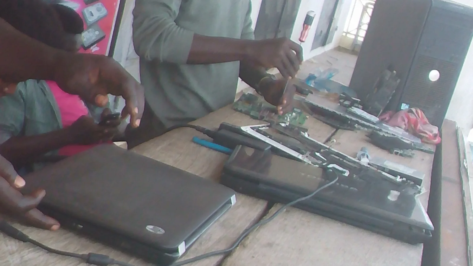 Two people at a wooden repair bench; one holds a closed laptop while the other uses a screwdriver on an opened laptop's exposed circuit board, with dismantled laptop chassis and a desktop tower on the table