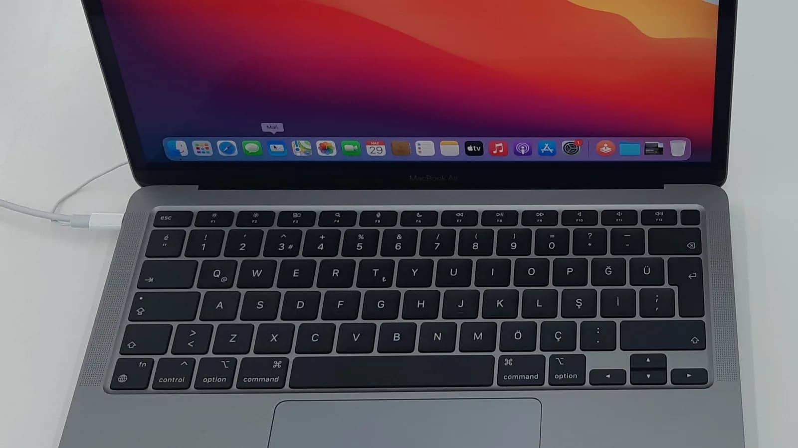 Sebuah komputer riba Apple berwarna kelabu angkasa yang terbuka di atas permukaan putih dengan kabel putih dipasang di sisi kirinya, skrinnya memaparkan kertas dinding merah dan jingga di atas sebaris ikon aplikasi, dengan papan kekunci dan trackpad kelihatan