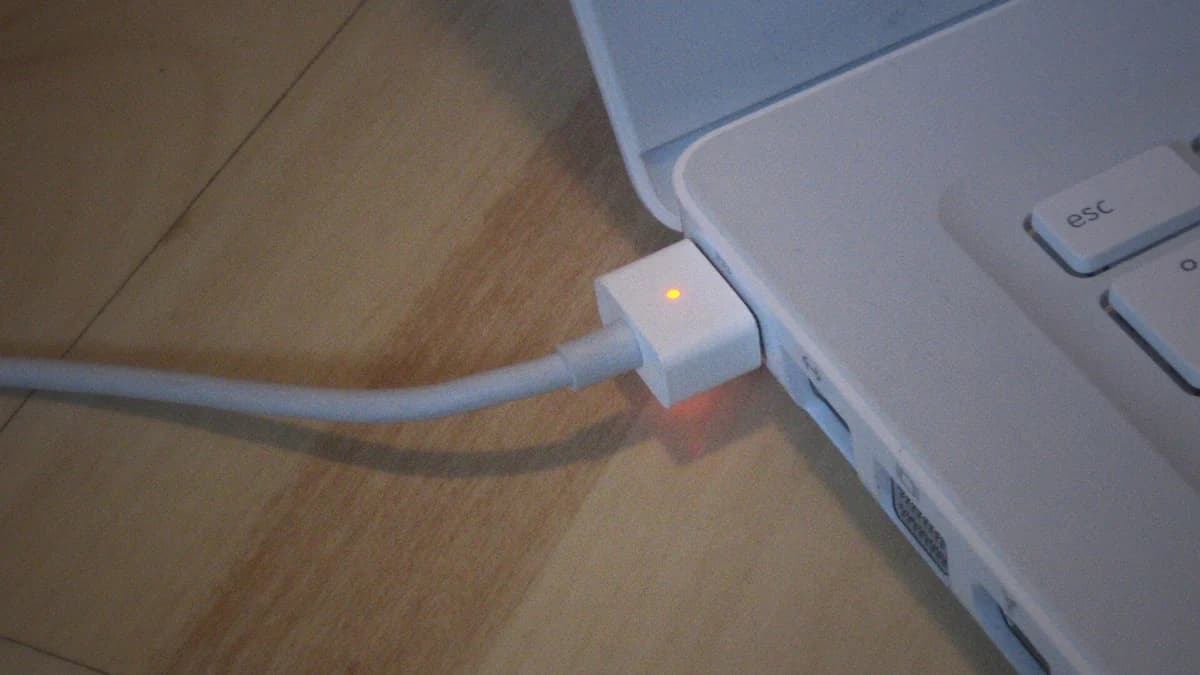 Penyambung kuasa MagSafe dipasang pada sisi sebuah MacBook, dengan lampu kuning kecil menyala di atas penyambung itu dan kabelnya melengkung di atas lantai kayu