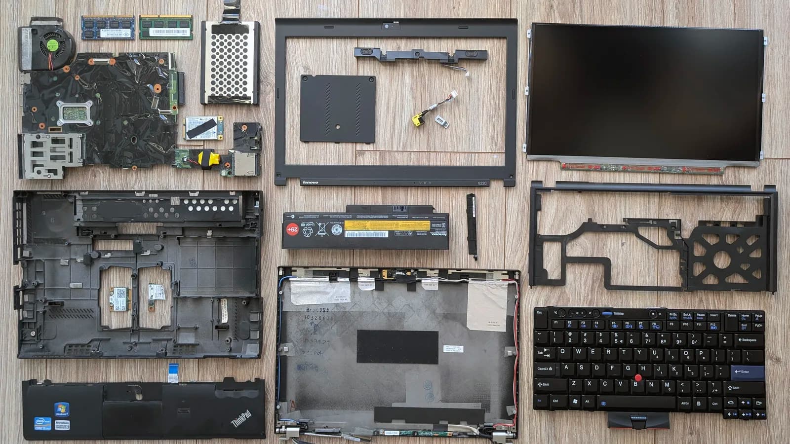 A laptop stripped completely apart and laid out in rows on a pale wooden surface: along the top a bare circuit board, metal brackets and small modules, with the bare dark display panel at the right; below them the empty black bottom case, the battery, a plastic frame piece and a black keyboard