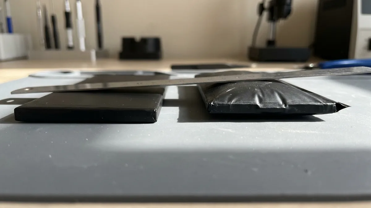 Two laptop battery packs side by side: the left one flat, the right one swollen so its centre balloons well above the bench, with a plain straight bar laid across both to show the gap.