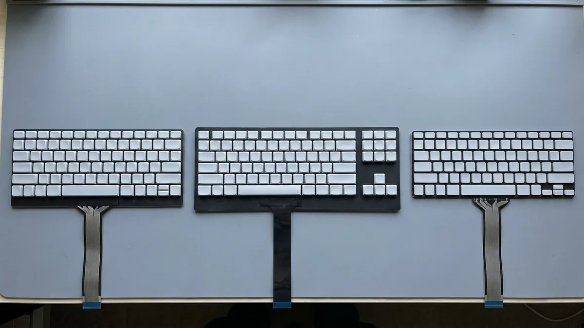 Three laptop keyboard modules of different widths and key layouts laid out in a row and seen from above, each on its own backing plate with a ribbon tail towards the camera.