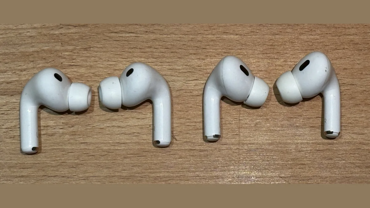 Four white AirPods Pro earbuds laid out in two pairs on a pale wooden surface, each with a white silicone ear tip and a small black port on the outer face