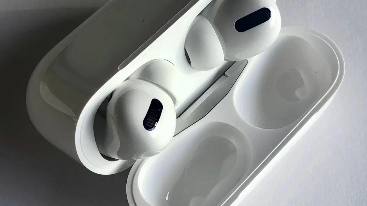 Two white AirPods Pro earbuds lying across their open charging case, the black oval speaker mesh clearly visible on each, with the two empty charging wells beside them