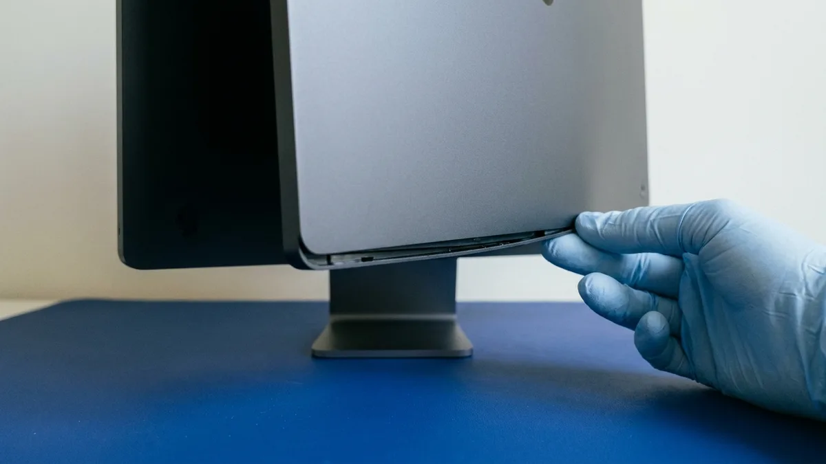 A pale blue gloved hand easing the plain rear cover of an all-in-one desktop computer a few millimetres clear along its lower edge, opening one fine dark gap, the machine standing on its pedestal foot on a deep blue anti-static mat.