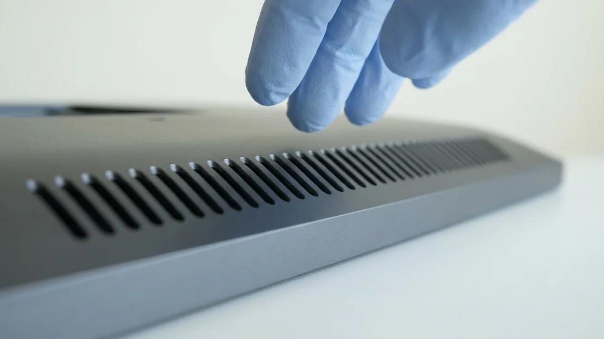 An extreme close-up of the fine slotted cooling vents running along the edge of a grey metal device, their narrow dark openings in a crisp even row, with pale blue gloved fingertips at the vent row.