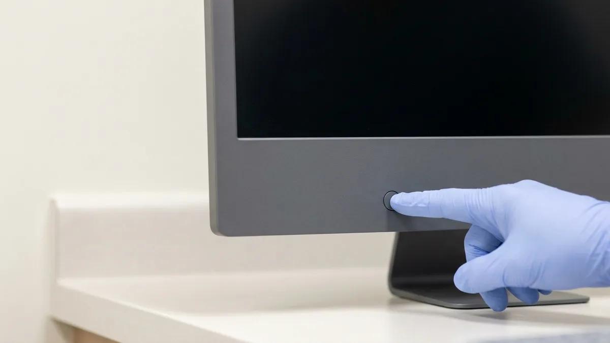 A pale blue gloved finger pressing the small plain round power button on the lower front edge of an all-in-one desktop computer standing on a spotless pale counter, its screen still dark and blank.