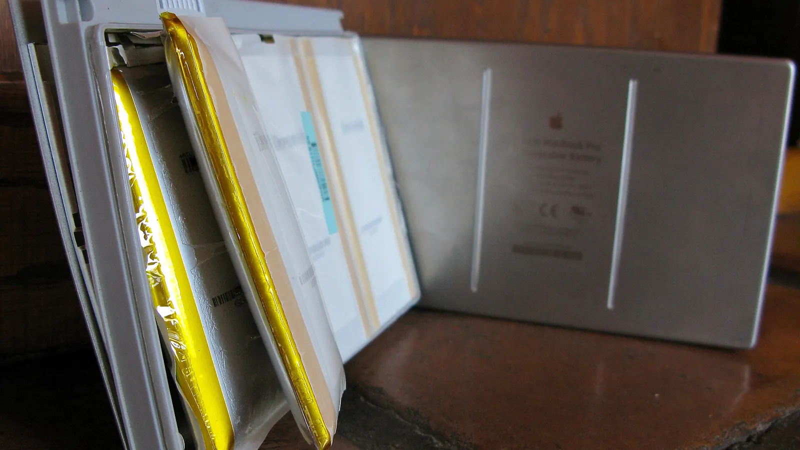 A white MacBook battery pack split open on a wooden ledge, its gold-foil pouch cells swollen so far that they have burst out of the plastic casing and no longer sit flat, with the intact back of the casing standing behind it