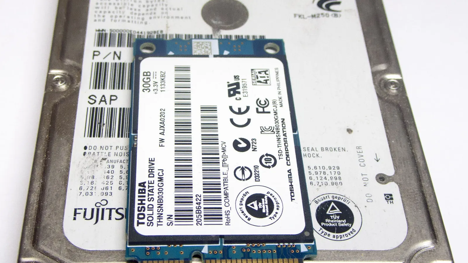 A small blue-green mSATA solid-state drive resting on top of a 2.5-inch laptop hard disk, both face up on a white surface so their printed labels, barcodes and certification marks are visible