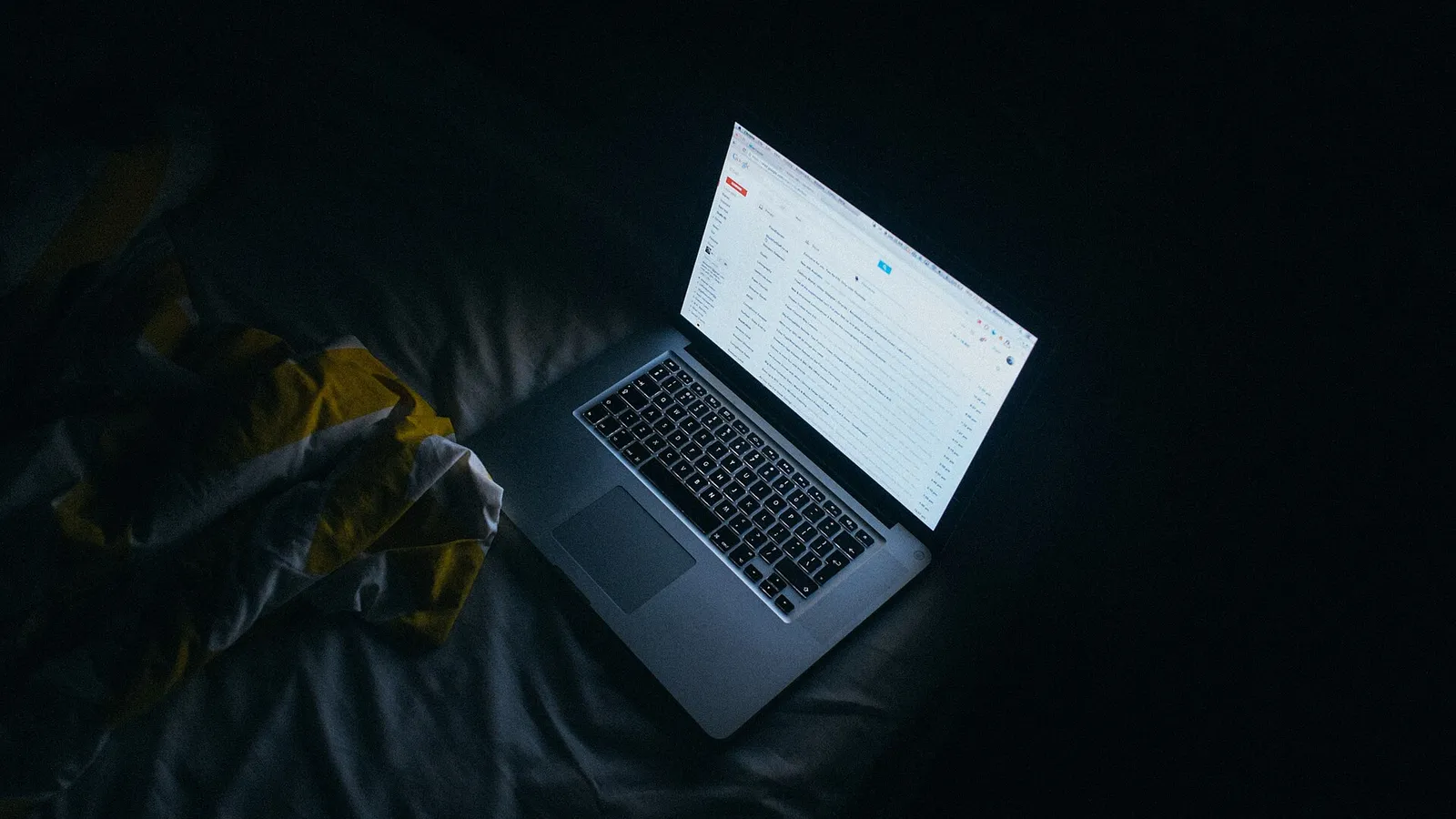 A MacBook open on a dark bed at night, its screen the only light in the frame, showing a list of email in an inbox, with a rumpled yellow and white sheet beside it