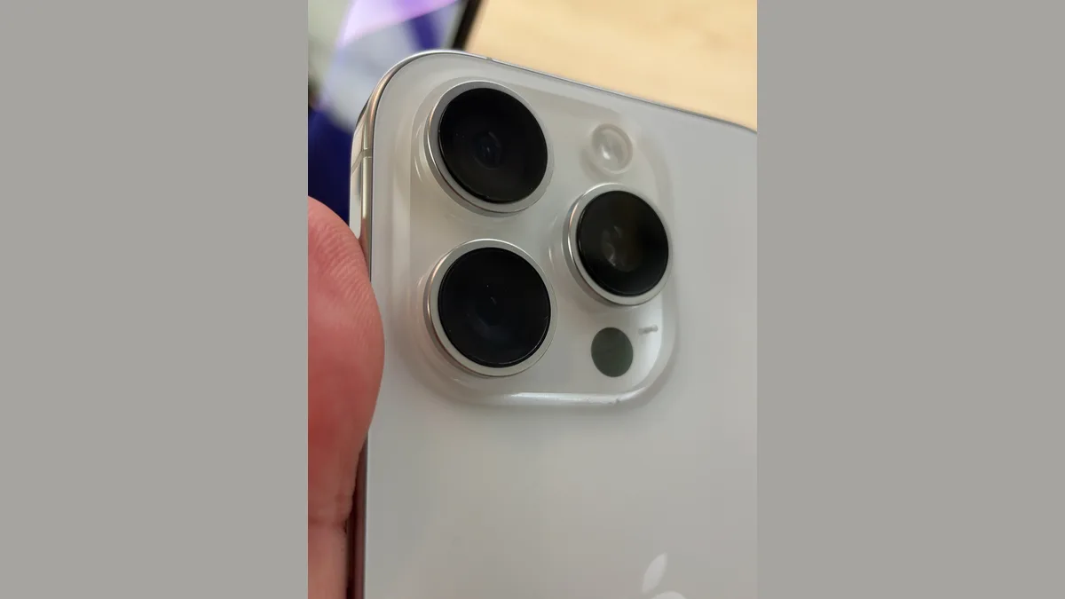 白色钛金属 iPhone 后置相机模块特写，三颗镜头嵌在凸起的金属环里，旁边是闪光灯和感应器小孔