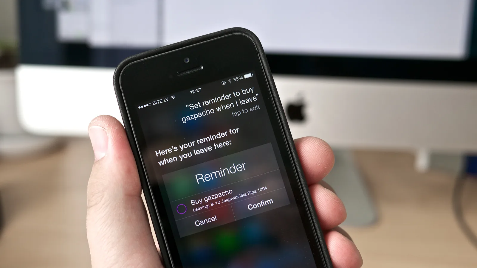 A hand holding a small older black iPhone in front of an iMac. On the screen Siri has transcribed the spoken request “Set reminder to buy gazpacho when I leave” and is offering a Reminder card with Cancel and Confirm buttons