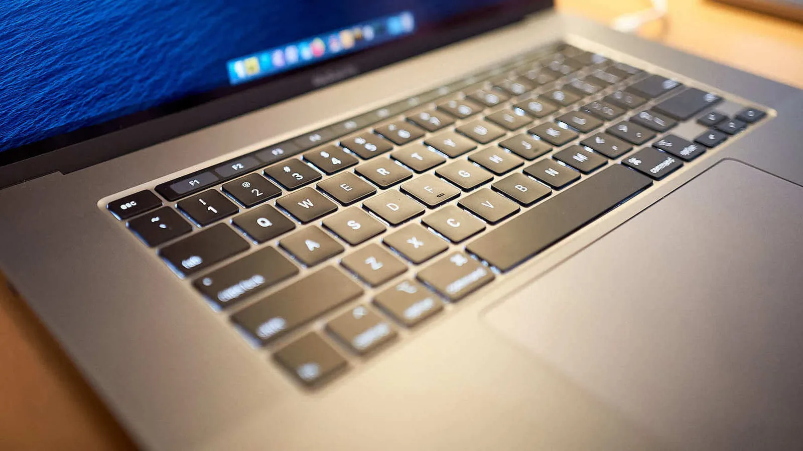 A 16-inch MacBook Pro open on a wooden desk, photographed low along the keyboard so the keys and the narrow Touch Bar strip above the top row are in focus, with a blue desktop wallpaper glowing behind