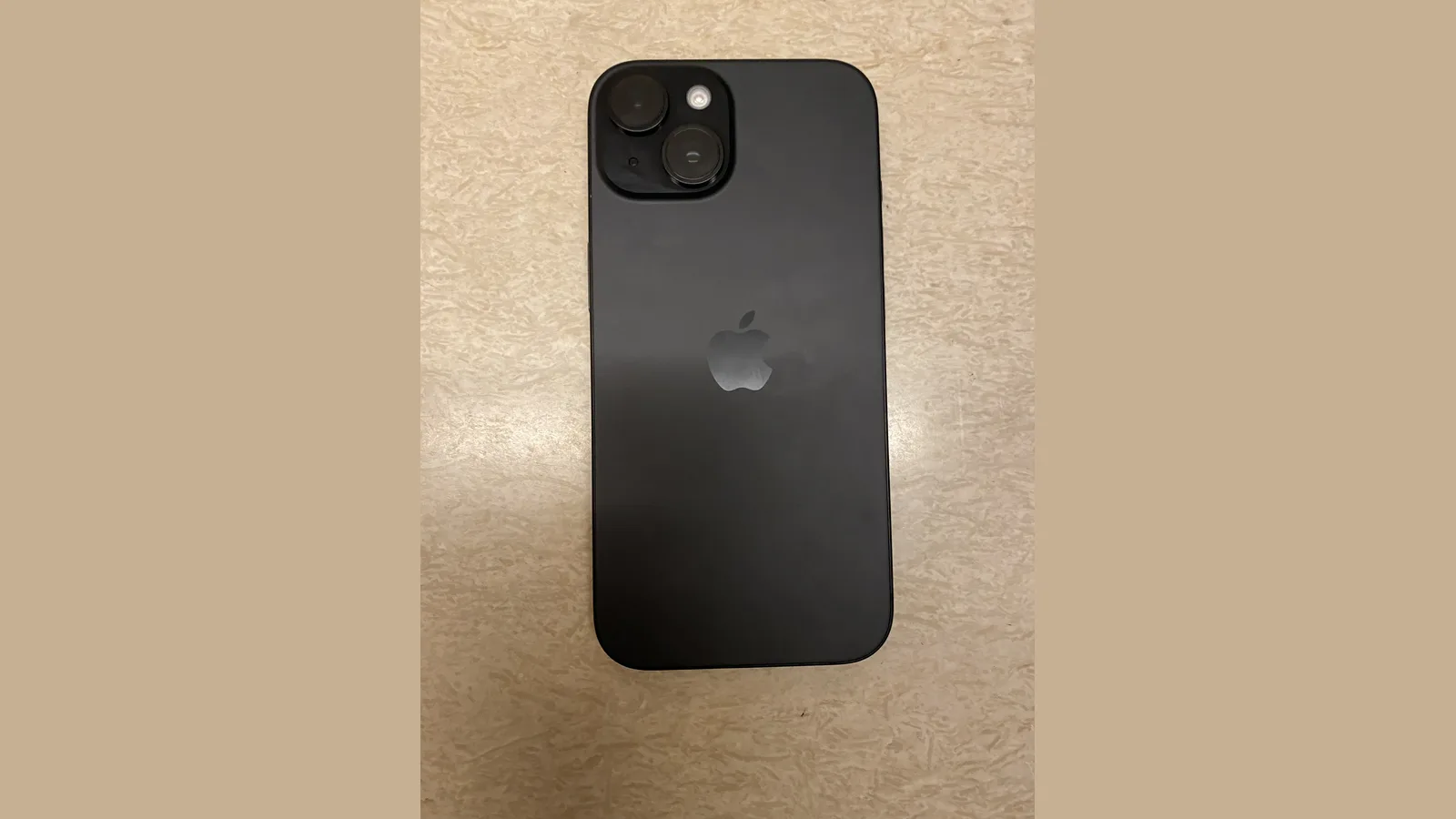 Bahagian belakang sebuah iPhone hitam dengan dua kanta kamera dan logo Apple, terletak di atas permukaan bercorak batu berwarna kuning air