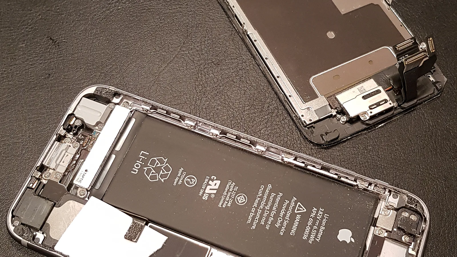 Sebuah iPhone dibuka untuk pembaikan di atas alas gelap: bateri Apple hitamnya dan jalur pelekat putih terdedah, dan pemasangan skrin terletak terlangkup di sebelahnya
