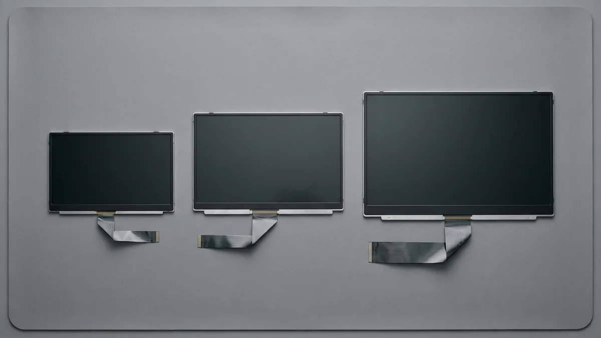 Three bare laptop display panels of clearly different sizes lie flat in a row on a plain grey mat, seen from directly overhead, each a thin rectangle with a dark unlit glass face and a flat ribbon cable of a different width folded out from its lower edge.