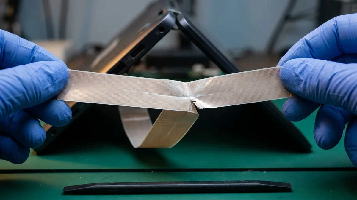 A wide pale flat ribbon cable held taut between two blue-gloved hands, a whitened worn crease running across its fold and a small frayed split opening at the edge of that crease, with the dark laptop lid it came from propped open behind and a black spudger lying flat on the green mat below.