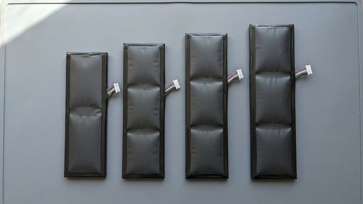 Four replacement laptop battery packs of different sizes laid out in a row and seen from above, each a set of flat pouch cells under a black wrap with a short wire bundle and plug.
