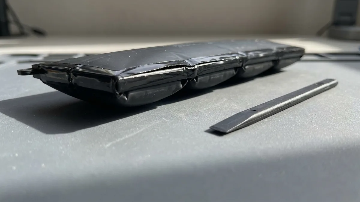 A close-up of a badly swollen laptop battery pack on a bench, its cells ballooned into rounded pillows that split the wrap along one seam and bow the pack clear of the surface beneath it.