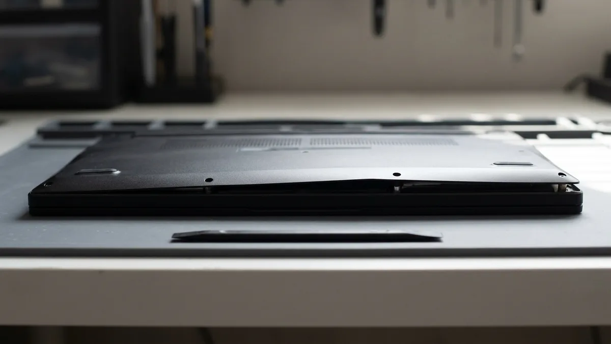 A laptop lying open-based on a bench where the swollen battery inside has bowed the chassis, a steel ruler laid across it showing daylight beneath its middle.