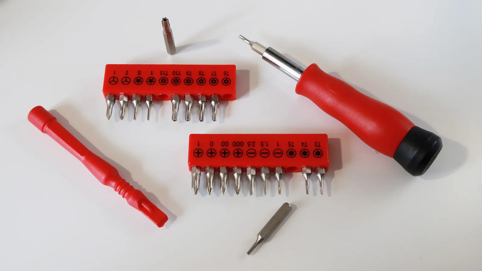 A red-handled precision screwdriver with two strips of bits for different screw heads, two loose bits and a red plastic prying tool on a white table.