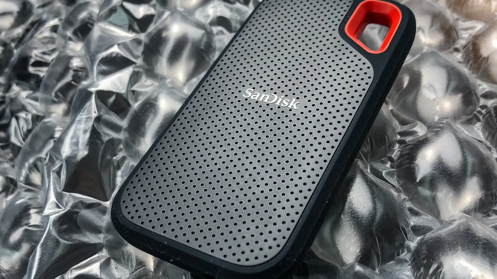 Sebuah SSD mudah alih SanDisk kelabu gelap yang padat dengan permukaan bertekstur bintik dan gelung sudut oren, terletak di atas permukaan perak berkedut