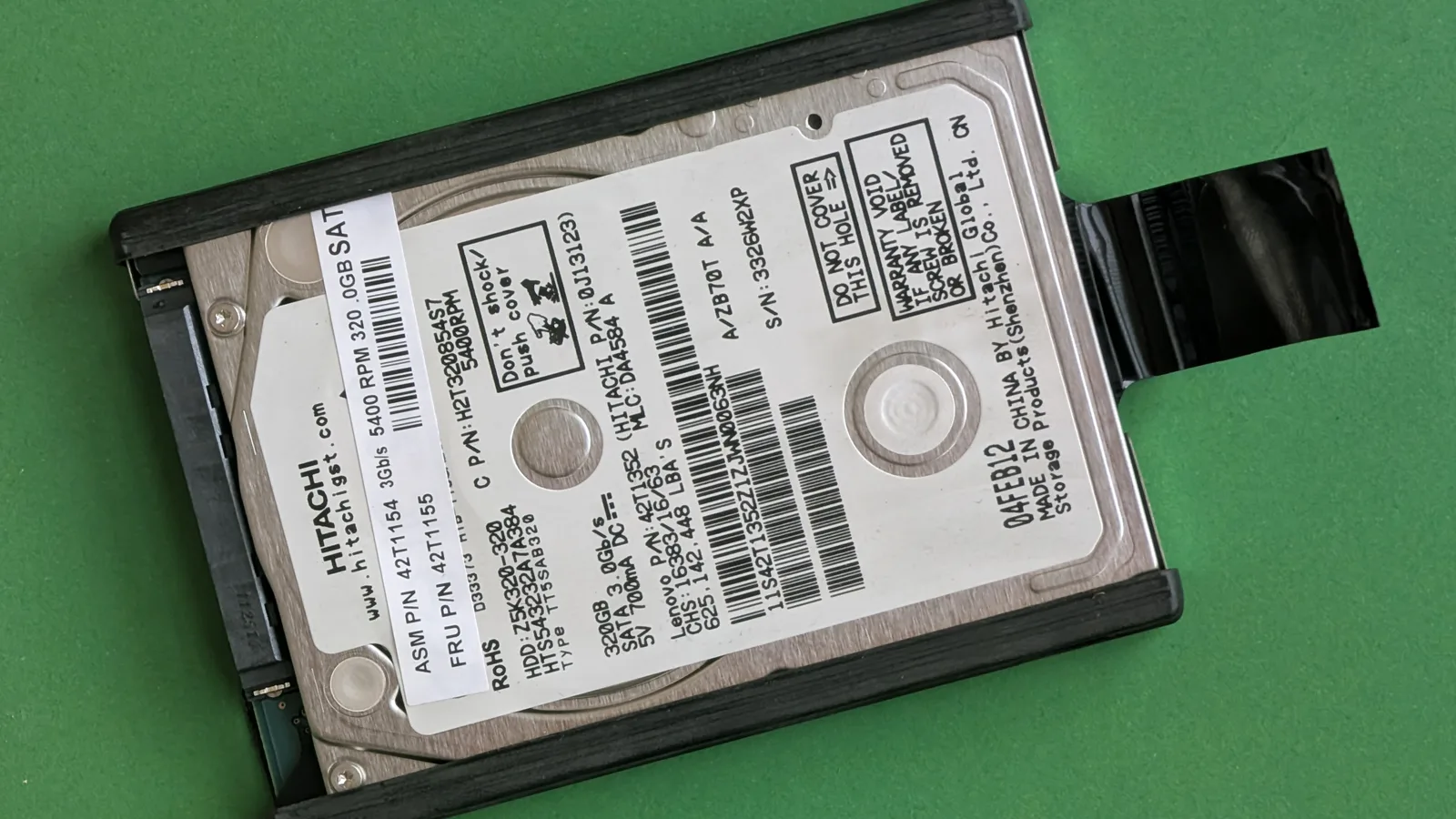 Pemacu keras komputer riba Hitachi 2.5 inci dengan label spesifikasi putih, dipasang dalam kadi pemacu hitam dengan penyambung pita, terbaring di atas permukaan hijau