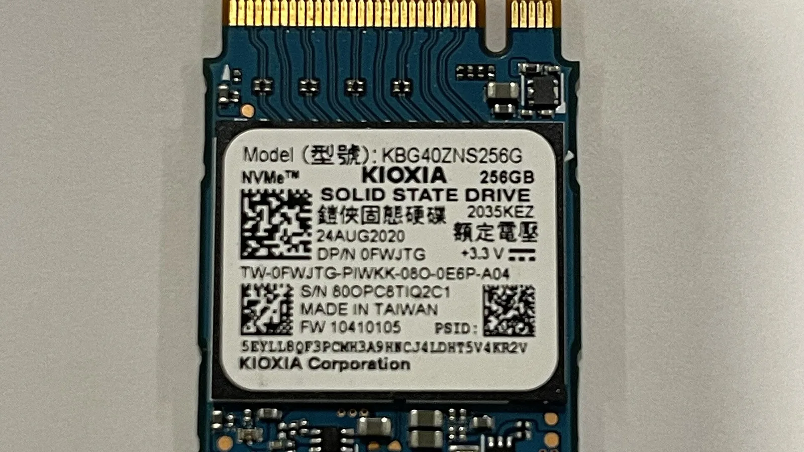 Pemacu keadaan pepejal M.2 NVMe kecil berwarna biru yang terletak rata di atas permukaan kelabu pucat, dirakam dari atas dengan pin sentuhan emasnya di sepanjang tepi atas; label putihnya tertulis 'KIOXIA', '256GB', 'SOLID STATE DRIVE' dan 'MADE IN TAIWAN', dengan tiga kod bar segi empat