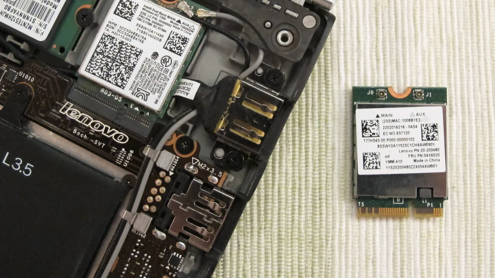Bahagian dalam komputer riba yang dibuka di sebelah kiri: papan induk gelap bercetak 'lenovo', sudut bateri hitam bertanda 'L3.5', wayar antena kelabu dan hitam serta kad tanpa wayar berlabel putih di dalam slotnya; di atas tikar berjalur krim di sebelah kanan terletak sekeping kad tanpa wayar hijau kecil yang kedua dengan label putih tertulis 'MAIN', 'AUX' dan 'Lenovo PN', dengan sesentuh emas di sepanjang tepi bawahnya
