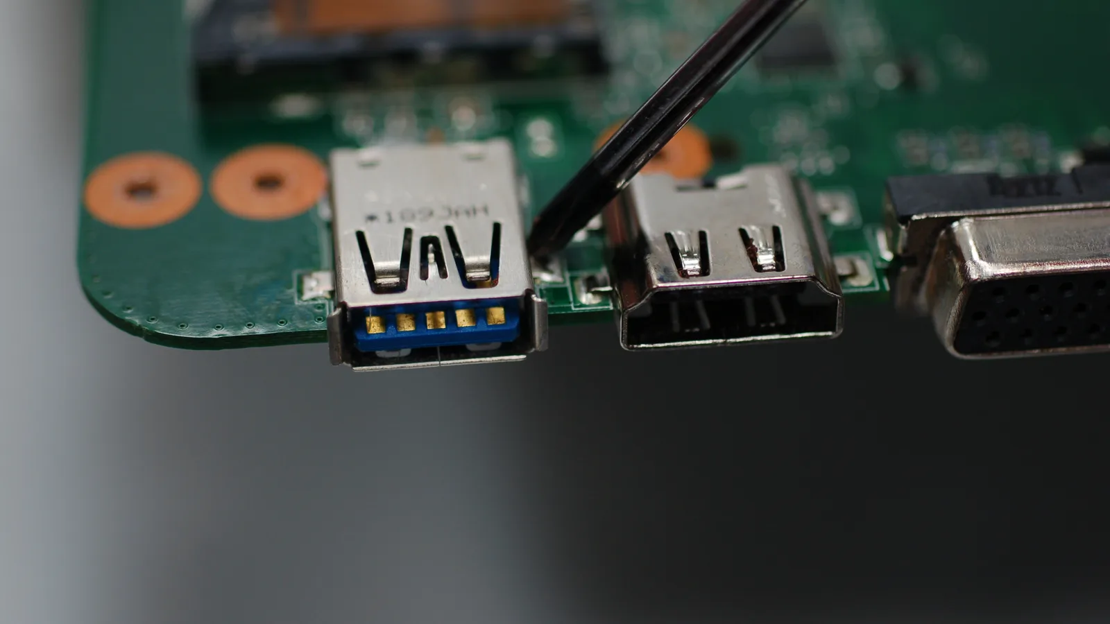 Close-up tepi papan induk komputer riba hijau menunjukkan port sisi yang dipateri padanya — port USB 3.0 biru, port HDMI dan penyambung VGA — dengan alat logam nipis bermata tirus menyentuh sambungan pateri di sebelah port USB
