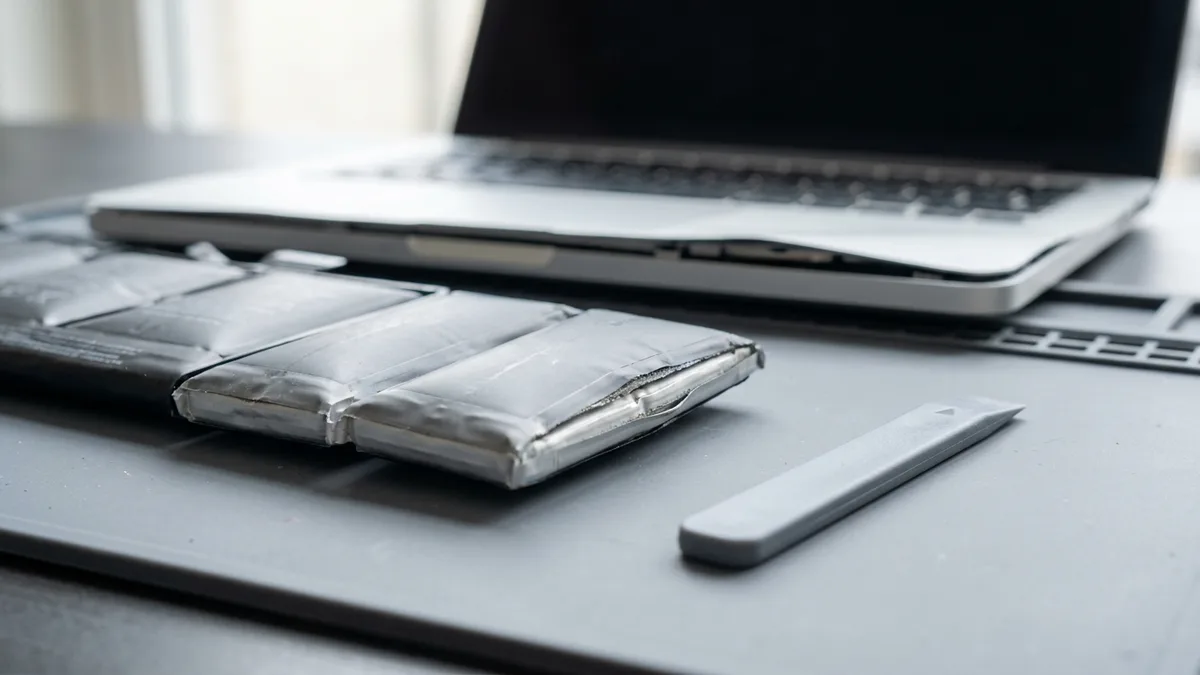 Swollen laptop battery pack on a workbench mat, its pouch cells ballooned and split along the seam, beside the opened laptop