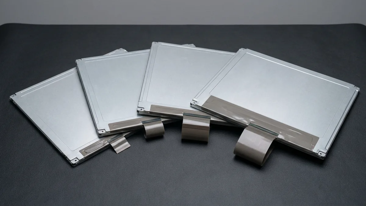 Four bare laptop display panels of different sizes lie fanned out in a row on a dark bench mat, every one face-down so only the plain silver metal rear chassis shows, each with a flat ribbon cable of a different width folded out along its lower edge.