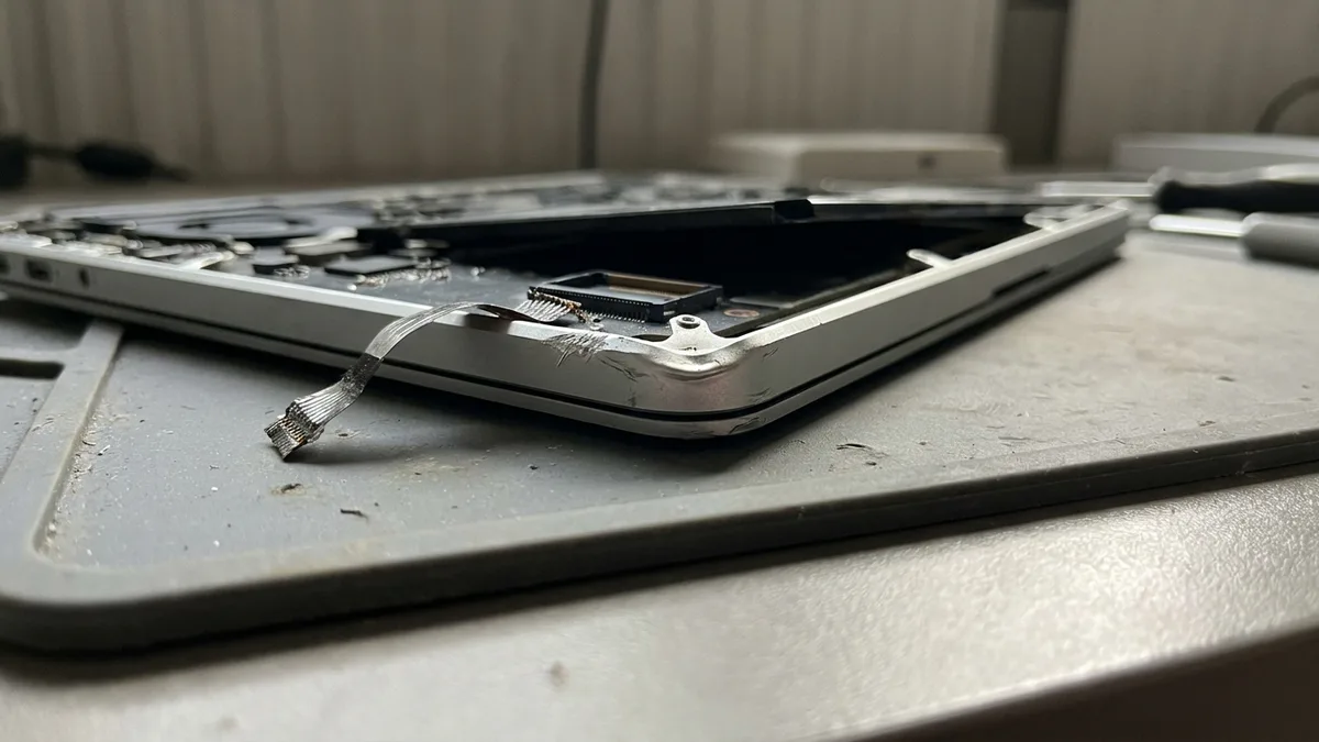 A close view of an opened silver laptop lower case on a bench mat, with a thin flat ribbon cable torn out of its small dark socket and lying curled loose, deep bright scratches gouged into the metal frame beside the socket and one corner of the frame lifted.