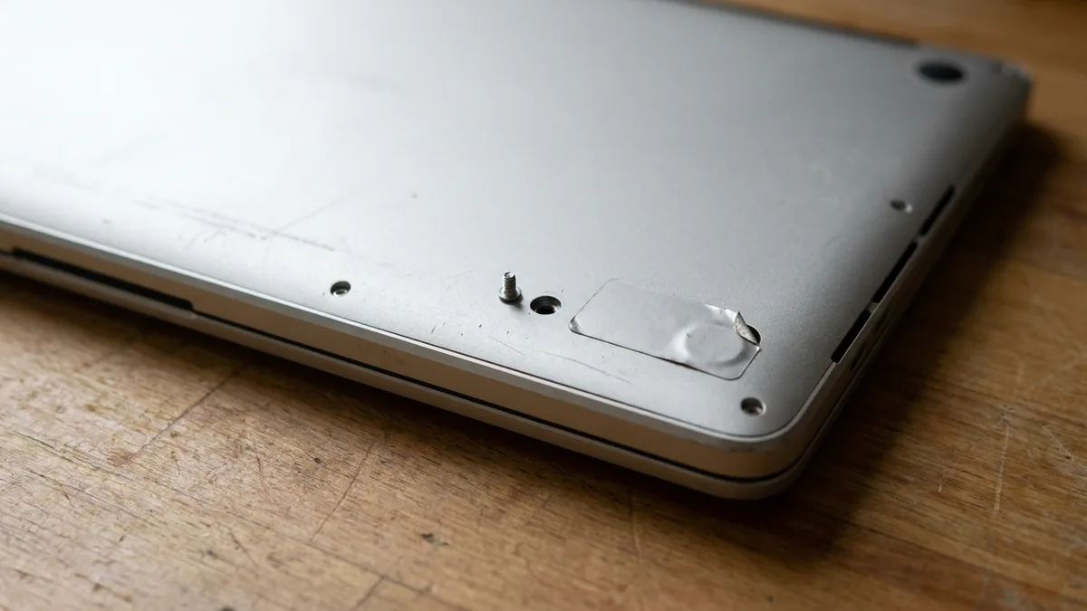 The smooth unmarked underside of a closed silver laptop on a pale counter, with one tiny screw sitting alone beside an empty screw hole and a small plain seal over a nearby hole split and peeling back at one corner.