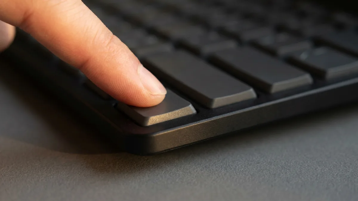 A fingertip pressing a single unmarked key near the bottom-left corner of a slim black keyboard, the key visibly pushed down below the keys beside it